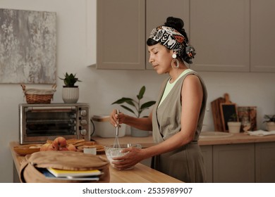 Woman mixing ingredients while standing in modern kitchen and showing dedication. Kitchen appliances and potted plants in the background providing a cozy atmosphere - Powered by Shutterstock