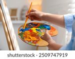 Woman mixing paints on palette with brush near easel indoors, closeup