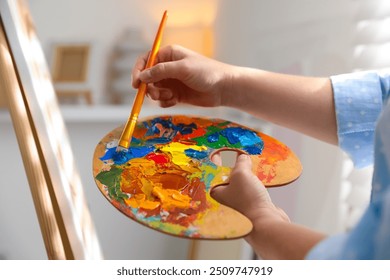 Woman mixing paints on palette with brush near easel indoors, closeup - Powered by Shutterstock