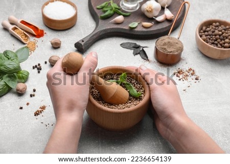 Woman mixing peppercorns and basil in mortar at light grey table, closeup