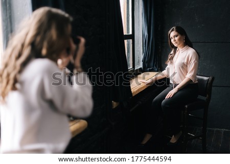 Woman model posing while sitting on chair in photo studio for unrecognizable female photographer which shooting with camera. Concept of creative work in photo studio, backstage job.