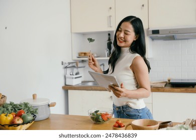 A woman in a modern kitchen preparing a meal, holding a digital tablet for recipe inspiration, surrounded by fresh vegetables. - Powered by Shutterstock