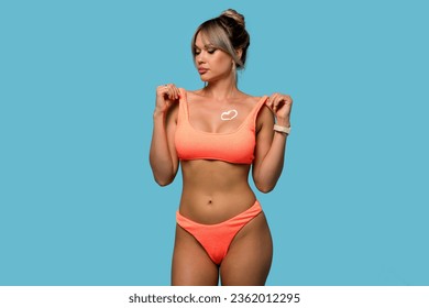 Woman with moisturizing sunscreen cream or body lotion on tanned breast in form of heart. Slender girl in orange swimwear on blue background. Perfect slim toned young body female