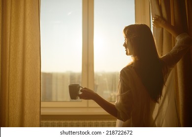 Woman In The Morning. Attractive Sexy Woman With Neat Body Is Holding A Cup With Hot Tea Or Coffee And Looking At The Sunrise Standing Near The Window In Her Home And Having A Perfect Cozy Morning.