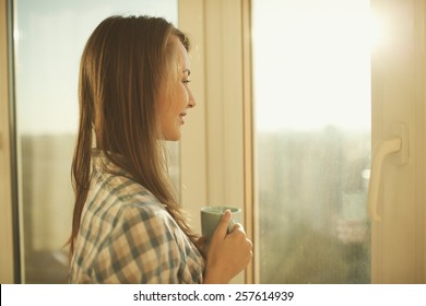 Woman in the morning. Attractive tender young woman is holding a cup with hot tea or coffee and looking at the sunrise standing near the window in her home and having a perfect cozy morning.
