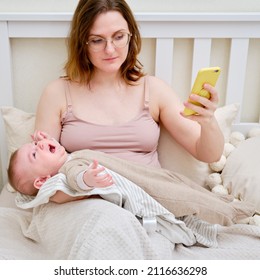 A Woman Mother Breastfeeding A Crying Infant Baby With A Phone In Her Hand. Mom Problems With Breastfeeding And Finding A Solution On The Internet