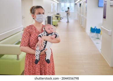 Woman Mother In Medical Face Mask Holds Baby Boy In Hospital Lobby. Mom With Child Son In Her Arms Visit In The Clinic Hall Waiting For A Doctor Appointment