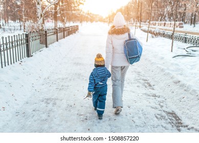 Woman Mother Walks Down Street Winter, With Small Child, Boy Or Girl 3-5 Years Old, View From Rear, Background Road Snowdrifts, Returns Kindergarten School Workout. Holiday Weekend Warm Clothes