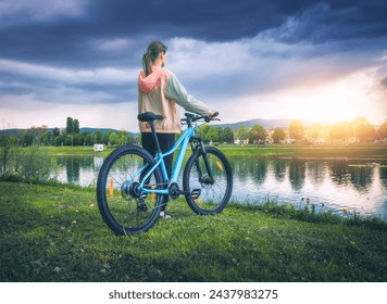 Woman with mountain bike near lake at overcast sunset in spring. Colorful landscape with sporty girl, bicycle, coast of river, green grass, cloudy sky in park in summer. Sport and travel. Biking - Powered by Shutterstock