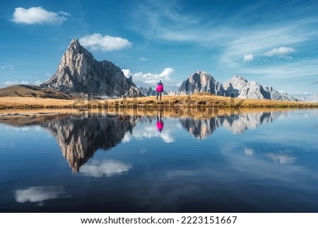 Woman and mountains reflected in lake at sunset in autumn in Dolomites, Italy. Standing girl on the shore of lake is looking on high rocks and blue sky with clouds in fall. Perfect reflection in water