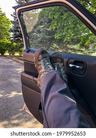 Woman In Muddy Hiking Boots And Work Pants Puts Feet Up On Car Door, Relaxing And Taking A Break In A Parking Lot Before Work