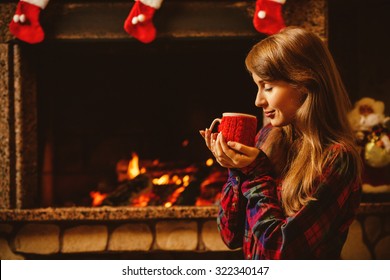 Woman With A Mug By The Fireplace. Young Attractive Woman Sitting By The Fireside And Holding A Cup With Hot Drink, Enjoying Cozy Evening. Holiday Time Concept In A House Decorated For Christmas.