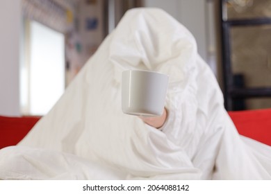 Woman with mug wrapped under the covers on couch cold weather, having a cold and lonely mood.