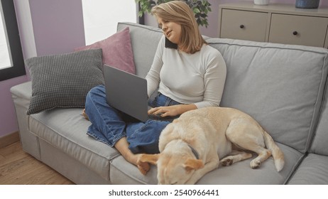 A woman multitasking at home, conversing on the phone while working on a laptop as her dog rests beside her on the couch. - Powered by Shutterstock