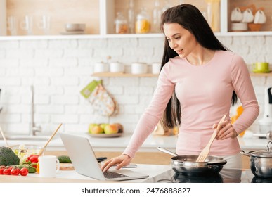 A woman is multitasking in the kitchen, cooking food while using a laptop for work or browsing the internet. She is standing near the stove, focusing on both tasks at hand. - Powered by Shutterstock