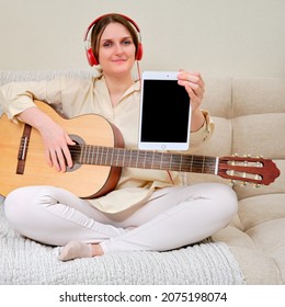 Woman musician with an acoustic guitar holds a tablet Apple iPad, mockup. Female guitarist sitting on a bed in a home living room with a tablet display, copy space - Moscow, Russia, November 01, 2021