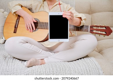 Woman musician with an acoustic guitar holds a tablet Apple iPad, mockup. Female guitarist sitting on a bed in a home living room with a tablet display, copy space - Moscow, Russia, November 01, 2021
