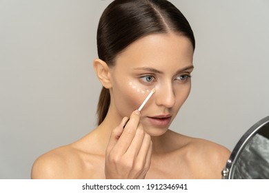 Woman with natural makeup applying concealer on flawless fresh skin, doing make up, looking at mirror. Girl after shower put corrector under eye area. Beauty face, skin care. Copy space, advertising.