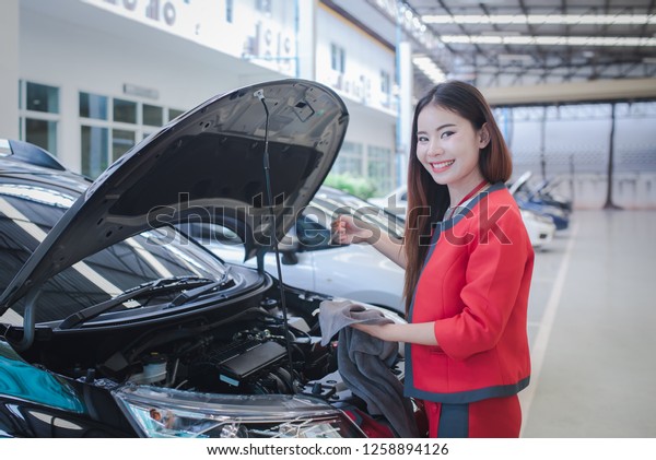 woman near car\'s hood. Beautiful Asian girl in\
covered parking of shopping center, stands near car with raised\
engine compartment hood, checks engine oil level in engine,\
inspects feeler gauge