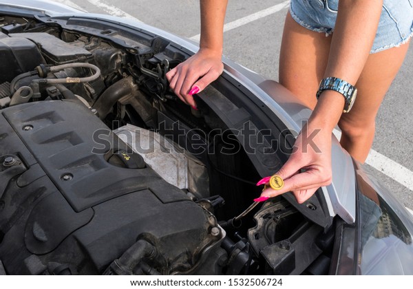 woman near car\'s hood.\
young girl in covered parking, stands near car with raised engine\
compartment hood, checks engine oil level in engine, inspects\
feeler gauge