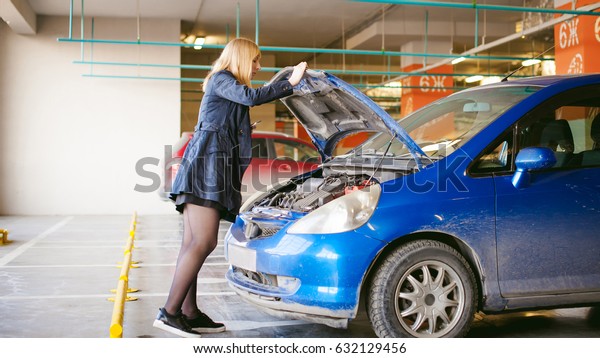 woman\
near car\'s hood. young blonde in covered parking of shopping\
center, stands near car with raised engine compartment hood, solves\
problem with malfunction by phone call to\
service
