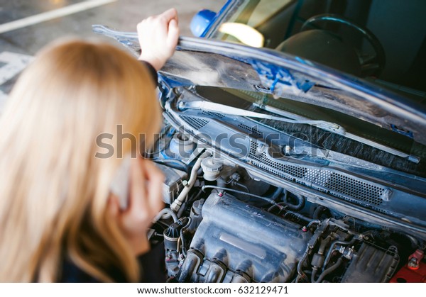woman\
near car\'s hood. young blonde in covered parking of shopping\
center, stands near car with raised engine compartment hood, solves\
problem with malfunction by phone call to\
service