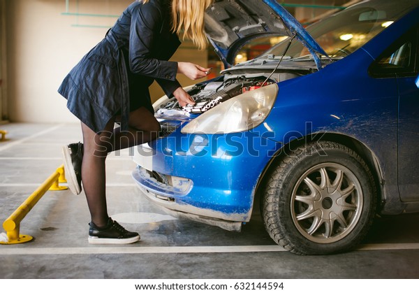 woman near car\'s hood. young blonde in covered\
parking of shopping center, stands near car with engine compartment\
bonnet raised, solves problem with engine malfunction, using\
carabiner wrench tool