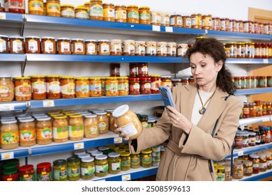 Woman near display of in Russian goods store. Buyer with jar of canned pickled fermented cabbage, scans QR code on product packaging, searches for information about manufacturer on Internet - Powered by Shutterstock