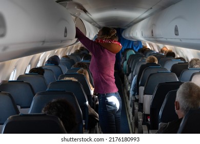 Woman With Neck Pillow Standing On Airplane Aisle Placing Suitcase Inside Overhead Compartment. Passenger Taking Carry On Luggage On Plane