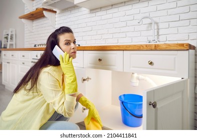 Woman Needs Help With Leaking Sink. Young Housewife Lady In Yellow Gloves Who's Trying To Stop Sink Pipe Leakage In The Kitchen Puts Blue Bucket Pail Under Drain And Calls Commercial Plumbing Service