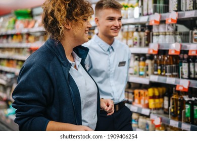 Woman With New Employee Walking In Supermarket. Male And Female Grocery Store Employees Talking And Smiling.