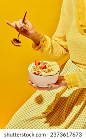 Woman in nice outfit holding golden spoon and bowl with muesli, cereal with berries against yellow background. Concept of healthy food, nutrition, pop art style, taste. Poster. Copy space for ad