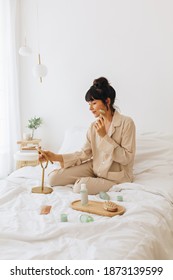 Woman In Night Suit Sitting On Bed At Home And Doing Routine Skin Care. Young Woman On Bed With Beauty Products And Mirror Doing Face Massage.