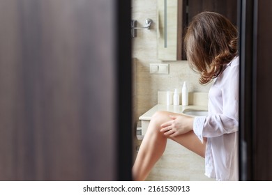 Woman In Nightgown Applying Cream For Protective And Care Dry Skin Legs Standing Against Mirror In Bathroom. 