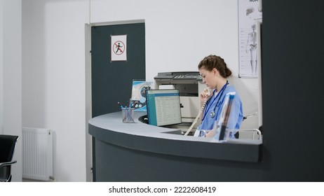 Woman nurse answering landline phone call at reception desk in healthcare facility, using cord telephone line to make medical appointments. Working at hospital registration counter. Handheld shot. - Powered by Shutterstock