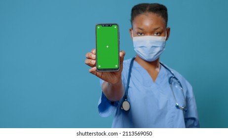 Woman Nurse With Face Mask Showing Green Screen On Smartphone In Studio. Healthcare Specialist Holding Mobile Phone With Chroma Key And Isolated Mockup Template During Covid 19 Pandemic.