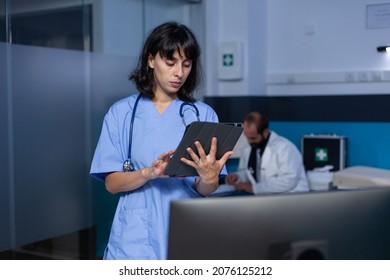Woman Nurse Looking At Digital Tablet Working Late At Night. Medical Assistant Holding Modern Gagdet With Touch Screen For Healthcare System And Medical Practice, Doing Work After Hours At Office.