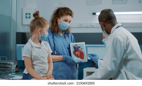 Woman Nurse Showing Cardiology Image On Digital Tablet, Letting Doctor Explain Cardiovascular Diagnosis To Child And Parent. Assistant Holding Modern Device With Heart Anatomy Picture.