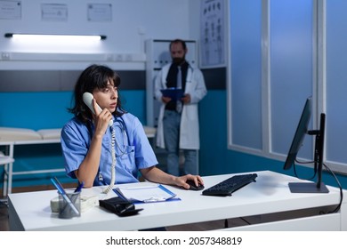 Woman Nurse In Uniform Using Landline Phone For Conversation With Patient About Healthcare Appointment. Medical Assistant Talking On Telephone And Looking At Computer, Working Late.