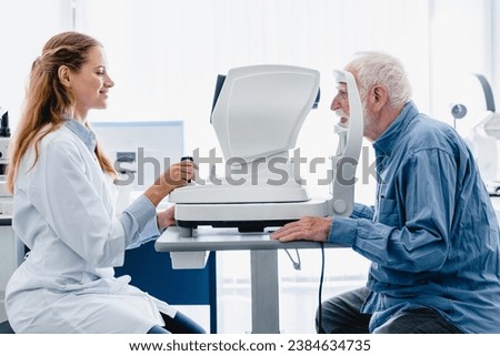 Woman oculist examining old man`s sight with ophthalmic tool in modern hospital clinic. Optician performing eyesight measurement for senior patient with myopia