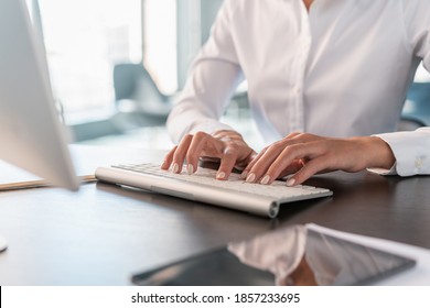 Woman office manager in white shirt sitting to a table with computer, hands on white keyboard closeup, no face. Female manicured nails typing on a keyboard, concept of office work