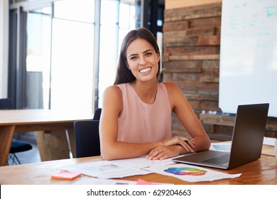 Woman in office using laptop smiles to camera, front view