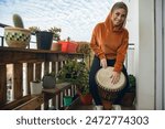 Woman on the balcony looking at the camera happy while playing her djembe drum. She is wearing an orange hoodie and is smiling. The balcony is filled with potted plants on recycled wood