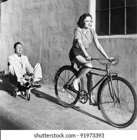 Woman on a bicycle pulling a grown man on a toy tricycle