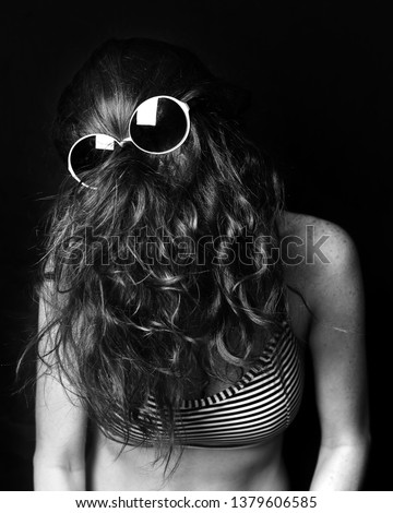 Woman on a black background with long thick wavy and messy hair covering or hiding her face, and disco glasses placed on the hair where her eyes would be, like a disguise, reminiscent of cousin itt.