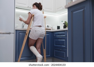 Woman On Crutches Washes The Dishes In The Kitchen. Temporary Limitation Of The Physical Capabilities Of A Person With A Broken Leg.