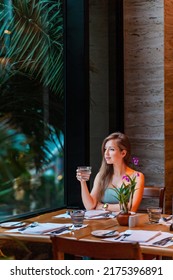 Woman On Dinner In Luxury Modern Restaurant. Attractive Girl Drinking Water From Glass, Looking At Window In Hotel Restaurant. Elegant Lady On Travel Vacation