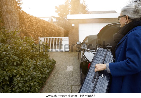 Woman on driveway in front of open car boot\
holding suitcase