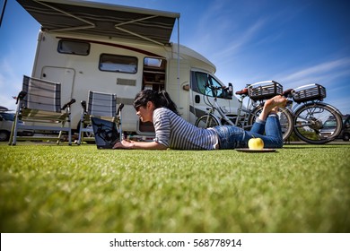 Woman On The Grass, Looking At The Laptop Near The Camping. Caravan Car Vacation. Family Vacation Travel, Holiday Trip In Motorhome