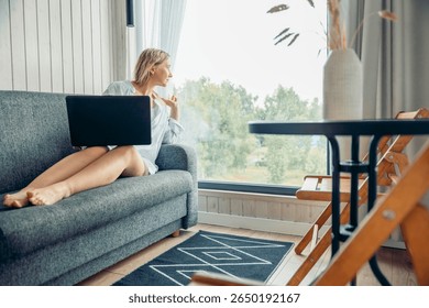 woman on gray sofa with laptop gazing thoughtfully out large window in bright room with wooden walls, remote work freelance coworking, business, e-commerce, online shopping